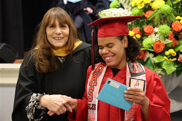 A Helen Keller School of Alabama graduate wears a red cap and gown while posing with a woman wearing black ceremonial robe and gold hood. They each smile excitedly. 